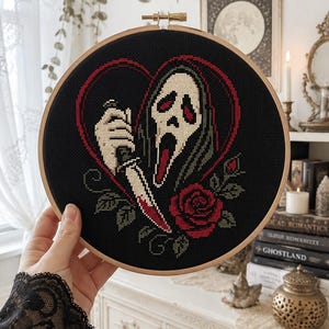 Può includere: Un telaio da ricamo circolare in legno contenente un disegno a punto croce su tessuto nero. Il disegno raffigura una maschera bianca spettrale con un cappuccio scuro, che impugna un coltello con la lama macchiata di rosso, incorniciato da un cuore e una rosa rossi.