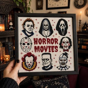 May include: A framed cross-stitch embroidery featuring the text HORROR MOVIES in red, surrounded by eight iconic horror character portraits including a hockey mask, a clown, and a nun, all accented with small red cross symbols on a cream fabric.