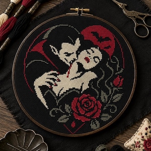 May include: A circular embroidery hoop containing a cross-stitch pattern of a vampire biting a person's neck. The design features a red moon, a bat silhouette, and red roses on a black background. Embroidery floss, scissors, and a pin cushion are nearby.