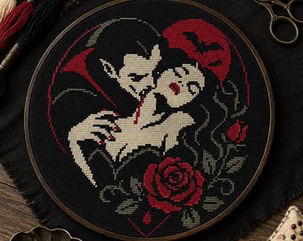 May include: A circular embroidery hoop containing a cross-stitch pattern of a vampire biting a person's neck. The design features a red moon, a bat silhouette, and red roses on a black background. Embroidery floss, scissors, and a pin cushion are nearby.