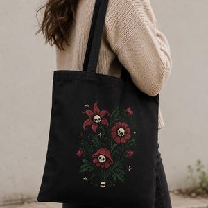 May include: A black tote bag featuring a cross-stitch style embroidery of dark red flowers with small white skulls at their centers, surrounded by green leaves and tiny white cross symbols, with two black shoulder straps.