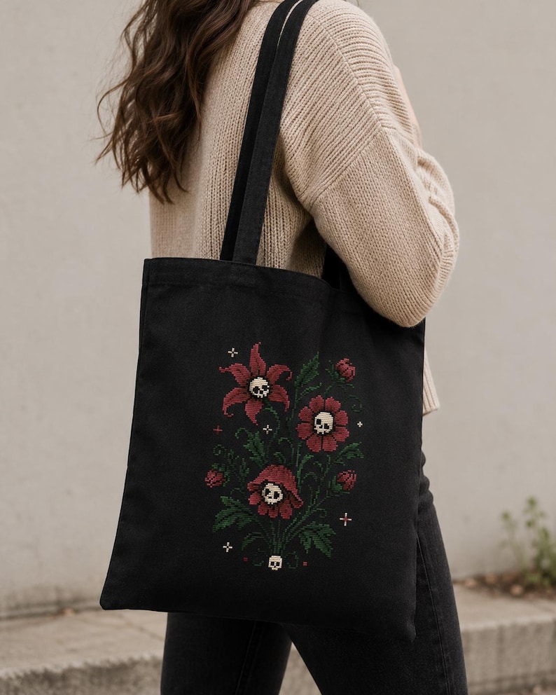 May include: A black tote bag featuring a cross-stitch style embroidery of dark red flowers with small white skulls at their centers, surrounded by green leaves and tiny white cross symbols, with two black shoulder straps.