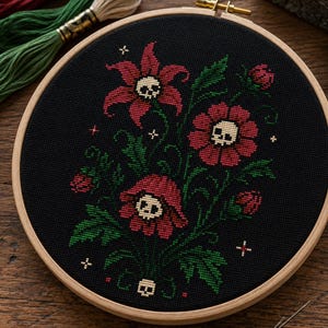 May include: A circular wooden embroidery hoop containing black fabric with a cross-stitch design of red flowers featuring small white skulls at their centers, surrounded by green leaves, stems, and tiny white cross-shaped accents.
