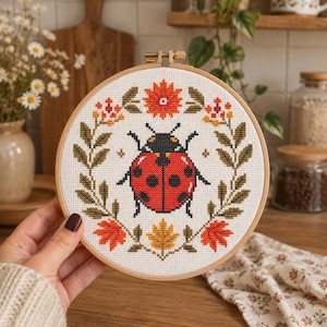 Peut inclure: Un tambour à broder circulaire en bois contenant un motif au point de croix représentant une coccinelle rouge à points noirs, centrée dans une couronne de feuilles aux couleurs automnales, de petites fleurs et une fleur orange au sommet.