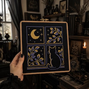 May include: A square cross-stitch embroidery piece in a wooden hoop featuring a four-pane window design on black fabric. The panels display a yellow crescent moon, purple and yellow leafy branches, and a silhouette of a cat sitting on a ledge.
