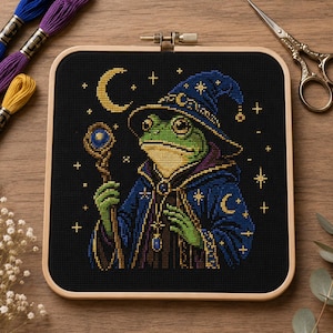 May include: Cross-stitch of a green frog wizard in a blue hat and robe, holding a staff. The design has a black background with gold stars and a crescent moon. The artwork is framed in a wooden hoop.