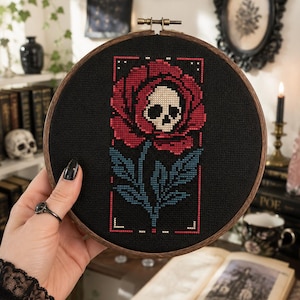 May include: A circular wooden embroidery hoop containing a cross-stitch design on black fabric. The design features a large red rose with a beige skull at its center, positioned above a blue stem and leaves, all enclosed within a thin red rectangular border.