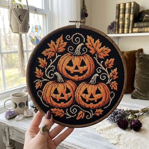 May include: A circular wooden embroidery hoop containing a cross-stitch design on black fabric. The design features three orange jack-o'-lantern pumpkins with carved faces, surrounded by orange autumn leaves and swirling vine patterns.