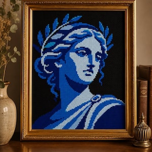 Op de afbeelding: Een ingelijst kruissteekkunstwerk met een blauw-witte afbeelding van een klassieke buste met een lauwerkrans. Het kunstwerk staat tegen een zwarte achtergrond en is ingelijst in een goudkleurige, sierlijke lijst.