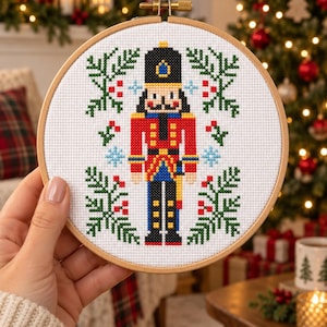 May include: A circular wooden embroidery hoop containing a cross-stitch pattern of a nutcracker soldier. The figure wears a black hat, red jacket, and blue trousers, flanked by green pine branches with red berries and light blue snowflake accents.