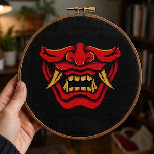 May include: A cross-stitch embroidery of a red and yellow Japanese demon mask on a black background. The mask is framed in a round wooden hoop. The design features sharp teeth and a fierce expression, perfect for wall decor.