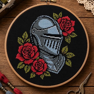 Può includere: Ricamo a punto croce in un telaio di legno con un elmo da cavaliere e rose rosse su sfondo nero. Il disegno è circondato da un telaio di legno. L'immagine è su una superficie di legno con filo da ricamo e forbici.