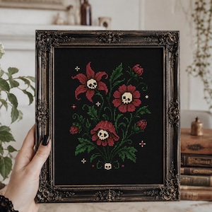 Könnte beinhalten: Eine gerahmte Kreuzstichstickerei auf schwarzem Stoff mit drei dunkelroten Blumen, die cremefarbene Totenköpfe in der Mitte haben, umgeben von grünen Blättern, kleinen Knospen und winzigen weißen Kreuzen, eingefasst in einem verzierten dunklen Rahmen.