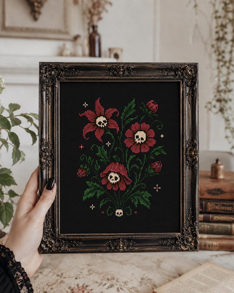 May include: A framed cross-stitch embroidery on black fabric featuring three dark red flowers with cream-colored skulls at their centers, surrounded by green leaves, small buds, and tiny white cross-shaped accents, all encased in an ornate dark frame.