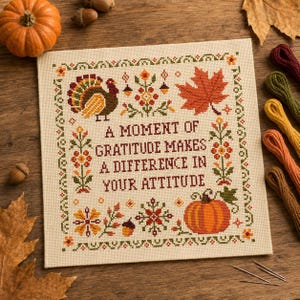 Puede incluir: Un muestrario de punto de cruz cuadrado sobre tela color crema con un pavo, una hoja de arce y una calabaza. El texto dice: A MOMENT OF GRATITUDE MAKES A DIFFERENCE IN YOUR ATTITUDE. El diseño está enmarcado por un borde decorativo de flores.