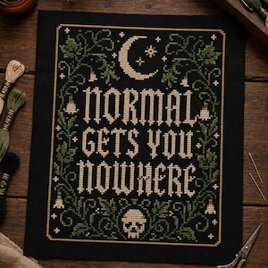 May include: A black cross-stitch embroidery on fabric featuring the text "NORMAL GETS YOU NOWHERE" in beige thread. The design includes a crescent moon, stars, a skull, and green vine borders with small flowers, all framed by a thin gold-coloured border.