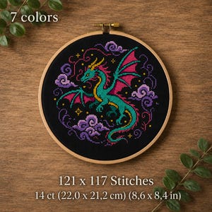 May include: Cross-stitch embroidery of a dragon with green body, pink wings, and yellow accents, surrounded by purple clouds, set against a black background. The text "7 colors", "121 x 117 Stitches", and "14 ct (8.6 x 8.4 in)" are visible.