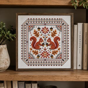 Puede incluir: Un bordado de punto de cruz cuadrado enmarcado en madera oscura, que muestra dos ardillas naranjas simétricas frente a frente sosteniendo bellotas. El diseño incluye hojas de roble, bellotas y motivos florales otoñales en tonos rojos, naranjas y marrones, todo encerrado en un borde geométrico sobre un fondo de tela blanca.