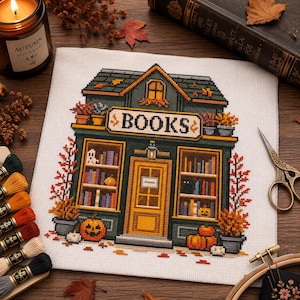 Pode incluir: Um padrão de ponto cruz em tecido branco representando uma livraria com exterior verde-escuro, uma porta amarela central e grandes vitrines cheias de livros coloridos. A loja apresenta a palavra BOOKS, abóboras e folhagem de outono.