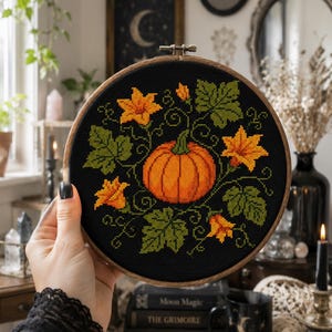 Op de afbeelding: Een ronde houten borduurring met een kruissteekontwerp op zwarte stof. Het ontwerp toont een centrale oranje pompoen omringd door groene bladeren, krullende ranken en geel-oranje bloemen in een cirkelvormig patroon.