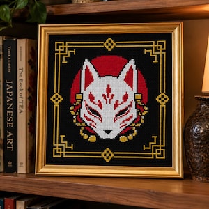 May include: A framed cross-stitch embroidery featuring a white and red Kitsune fox mask centered against a solid black background. The design is enclosed within a gold-colored geometric border and housed in a metallic gold frame.