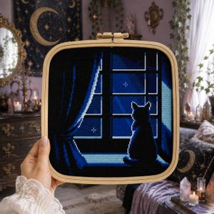Op de afbeelding: Een vierkant houten borduurraam met een kruissteekwerkje dat het silhouet van een kat op een vensterbank in de nacht toont. De afbeelding bevat een donkerblauw raamkozijn, gordijnen en een nachtelijke hemel met kleine witte sterren.