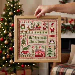 May include: A square cross-stitch sampler in a light wood frame featuring the text Merry Christmas. The design includes colourful embroidered motifs of a reindeer, a sleigh, Santa Claus, an owl, a red car, a house, and various evergreen trees.