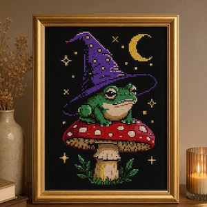 Puede incluir: Arte de punto de cruz de una rana verde con un sombrero de mago morado, sentada sobre una seta roja con lunares blancos. La obra enmarcada está sobre un fondo negro con una luna creciente y estrellas. El marco es dorado.
