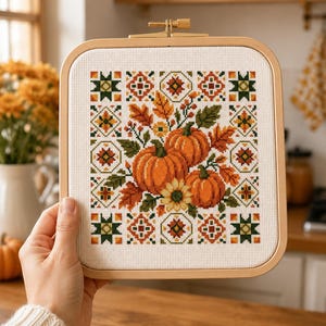 May include: A square wooden embroidery hoop containing a cross-stitch pattern on white fabric. The design features two orange pumpkins, autumn leaves, and a sunflower, surrounded by a geometric border of small green, orange, and brown motifs.