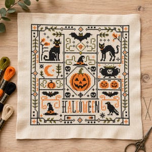 May include: A square piece of cream-colored Aida fabric featuring a detailed Halloween-themed cross-stitch pattern. The design includes black cats, bats, a jack-o'-lantern, a cauldron, skulls, and the word HALLOWEEN in black and orange thread.