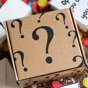 Puede incluir: Una caja de cartón marrón con signos de interrogación negros. La caja está rodeada de confeti colorido y una etiqueta que dice "Open if you dare!"