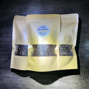 Pode incluir: Três sacos bege com uma janela transparente mostrando uma substância granular escura. Um adesivo azul na frente do saco central diz "LOVE YOUR ORDER? Please Leave a Review!"