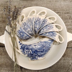 Peut inclure: Ensemble de tasses à thé et saucière en porcelaine bleue et blanche, disposées dans un plat de service blanc. Les tasses sont empilées en cercle. Une branche de lavande est placée sur le côté. La porcelaine présente un motif de paysage.