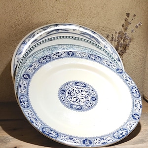 Peut inclure: Plusieurs assiettes en céramique blanche ornées de motifs bleus détaillés. Chaque assiette présente un motif central en médaillon et une bordure avec des figures et des motifs floraux. Les assiettes sont empilées, révélant les différents motifs.