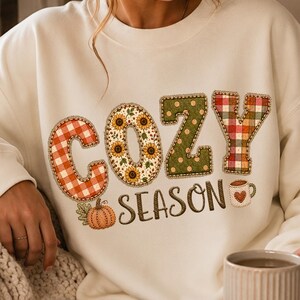 Peut inclure: Un sweat-shirt à col rond couleur crème avec le texte COZY SEASON dans une police décorative. Les lettres sont remplies de motifs incluant du vichy rouge, des tournesols, des pois verts et du tartan. Une petite citrouille orange et une tasse.