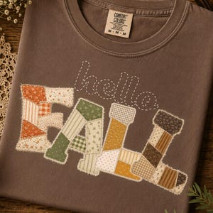Peut inclure: Un t-shirt marron avec le texte "hello" en contour blanc pointillé et le mot "FALL" en motif patchwork. Les lettres sont composées de divers motifs de tissu dans des tons orange, vert, jaune et marron avec des bords effilochés.