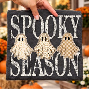 Può includere: Una t-shirt grigio scuro con la scritta bianca dall'aspetto consumato SPOOKY SEASON. Sotto la parola SPOOKY ci sono tre illustrazioni di fantasmi con motivi beige: uno a pois, uno con stampa leopardata e uno a scacchi.