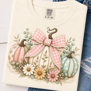 May include: A cream-colored t-shirt featuring a graphic print of three pumpkins decorated with a large pink and white gingham bow, lace trim, and various flowers including daisies. The shirt has a Comfort Colors brand label at the neck.