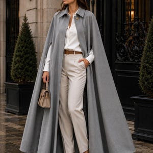 May include: A floor-length grey wool cape featuring a single gold-tone button closure at the collar, worn over a white long-sleeve button-down shirt, beige tailored trousers with a brown leather belt, and beige pointed-toe high-heeled pumps.