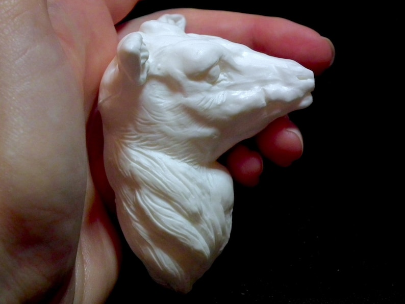 Op de afbeelding: Witte keramische sculptuur van een dierenkop, mogelijk een wolf of een hond, met gedetailleerde vachttextuur. De sculptuur wordt in een hand gehouden.