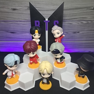 Può includere: Una collezione di sette figurine in miniatura di un gruppo musicale, ognuna con acconciature e abiti unici, esposte su una piattaforma esagonale bianca. Il logo BTS è visibile sullo sfondo. Le figurine sono su basi circolari gialle.