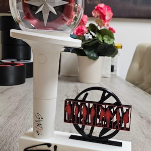 Può includere: Un bastoncino luminoso bianco e rosso con un disegno a stella all'interno di un globo trasparente. La base presenta il testo "Stray Kids" in scrittura nera e un logo nero e rosso. Un oggetto decorativo.