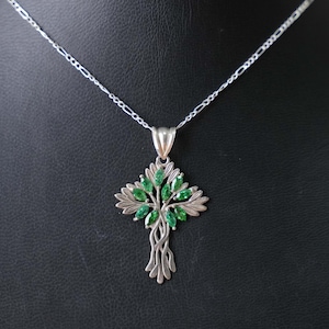 May include: Silver-toned pendant necklace featuring a tree design. The pendant has a detailed tree with green gemstone leaves. The necklace is displayed against a black background.