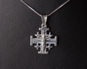 May include: A silver pendant necklace featuring a Jerusalem cross design. The pendant has a detailed, textured appearance with a figure of Jesus Christ at the center. The necklace chain is a delicate, silver box chain.