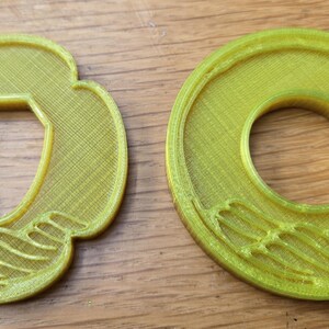 May include: Two yellow 3D-printed plastic objects with a textured surface. The object on the left has a clover-like shape with a central hexagonal cutout and wave-like ridges at the bottom. The object on the right is circular with a round centre hole.
