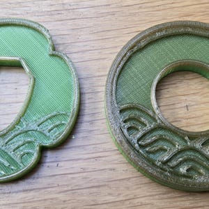 May include: Two 3D printed tsuba sword guards in a translucent green material. One guard is circular with a central hole, and the other is a four-lobed shape with a central opening. Both feature a raised wave pattern along the bottom edge.