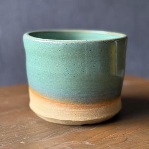 May include: A handmade ceramic tumbler with a turquoise glaze, transitioning to a tan base with an orange accent. The cylindrical cup is unadorned, showcasing the texture of the pottery. The cup is sitting on a wooden surface.