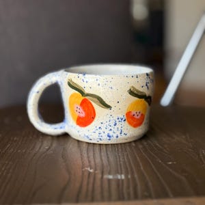 May include: A ceramic mug with a cream-colored base, adorned with hand-painted orange and red fruit designs and green leaves. The mug has a blue speckled pattern and a curved handle. The mug is sitting on a wooden surface.