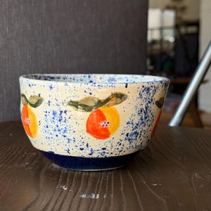 Puede incluir: Un cuenco de cerámica con exterior color crema, decorado con diseños de frutas naranjas y amarillas y motas azules. La base es azul oscuro y el cuenco está sobre una superficie de madera.