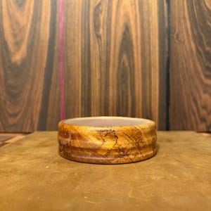 May include: A handcrafted wooden bowl with a natural, warm color palette. The bowl features a rounded shape with visible wood grain and a textured surface. The bowl is set on a brown surface, with a dark wood background.
